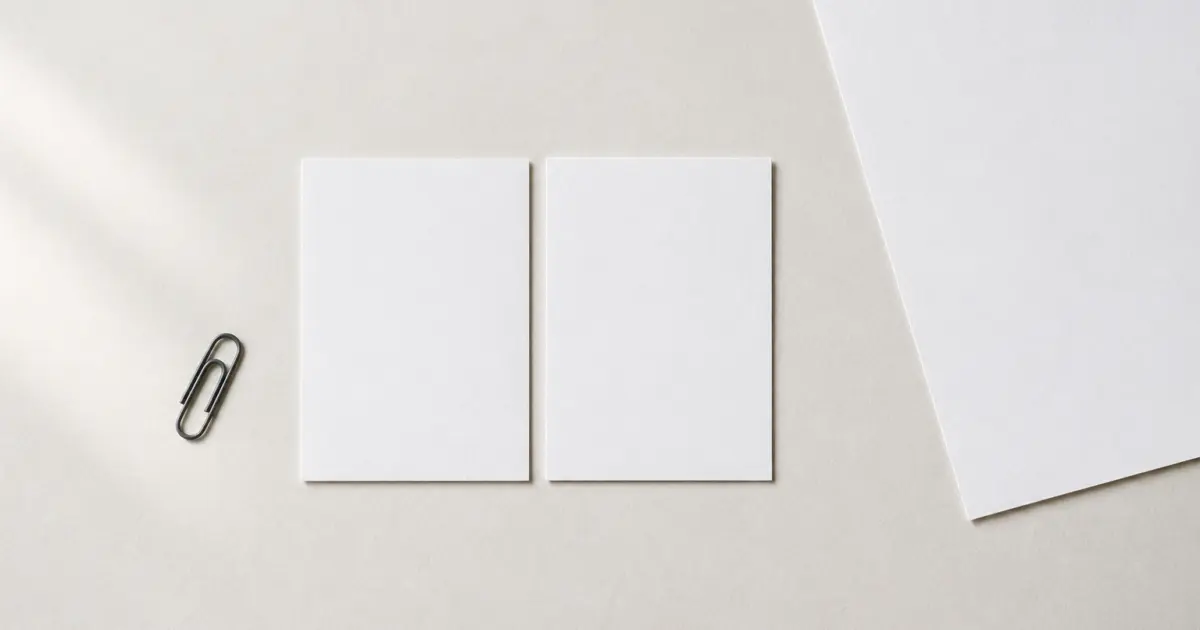 Two identical blank white cards squared up side by side on a pale desk, with a larger mismatched sheet pushed aside